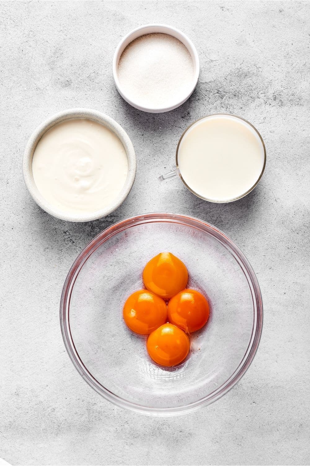 A glass bowls for a yolks in it, a cup of milk, a bowl filled with coconut cream, and a bowl filled with erythritol all on a gray counter.
