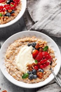 A white bowl filled with oatmeal with cream cheese, blueberries, strawberries, red currants, and pecans in the middle. Part of another bowl with the same thing is behind it.