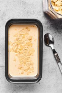 A loaf pan filled with almond milk ice cream with some sliced almonds on top. Next to it is a spoon and in front of that a small part of a glass jar filled with sliced almonds.