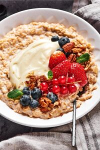 Blueberries, strawberries, red currants, pecans and cream cheese on top of oatmeal in a white bowl. A spoon is submerged in the oatmeal.