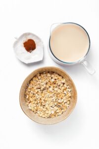 A bowl of rolled oats, a pitcher of milk, and a dish of cinnamon on a white counter.,