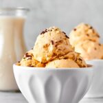 White bowl with a couple scoops of almond milk ice cream in it. Behind it is part of another ball scoops of ice cream and part of a glass jar with almond milk.