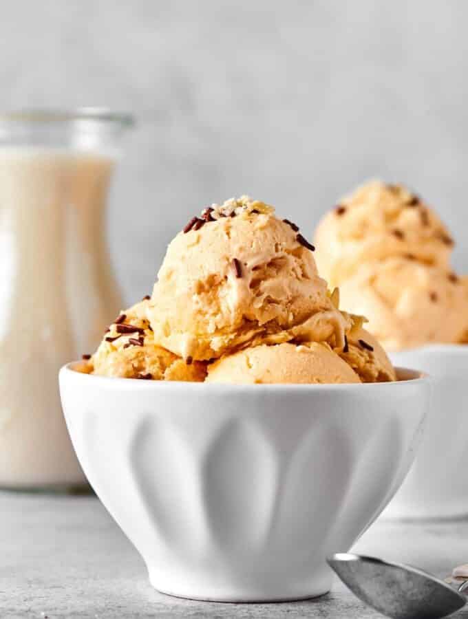 White bowl with a couple scoops of almond milk ice cream in it. Behind it is part of another ball scoops of ice cream and part of a glass jar with almond milk.