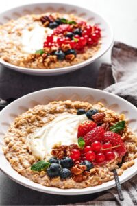 A bowl of oatmeal with cream cheese, blueberries, strawberries, red currants, and pecans in the middle. Another bowl filled with the same thing is behind it.