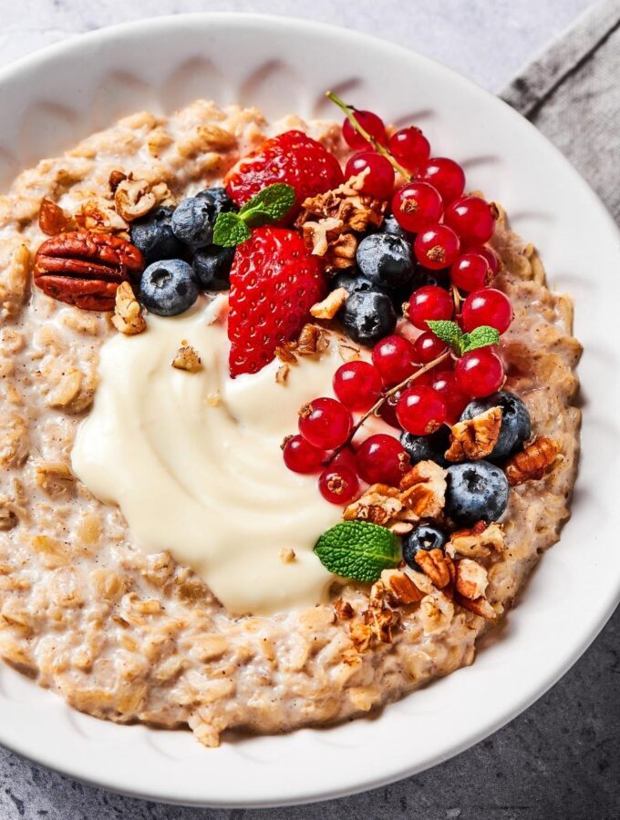 Oatmeal in a white bowl with cream cheese, blueberries, strawberries, red currants, and pecans on top of it.