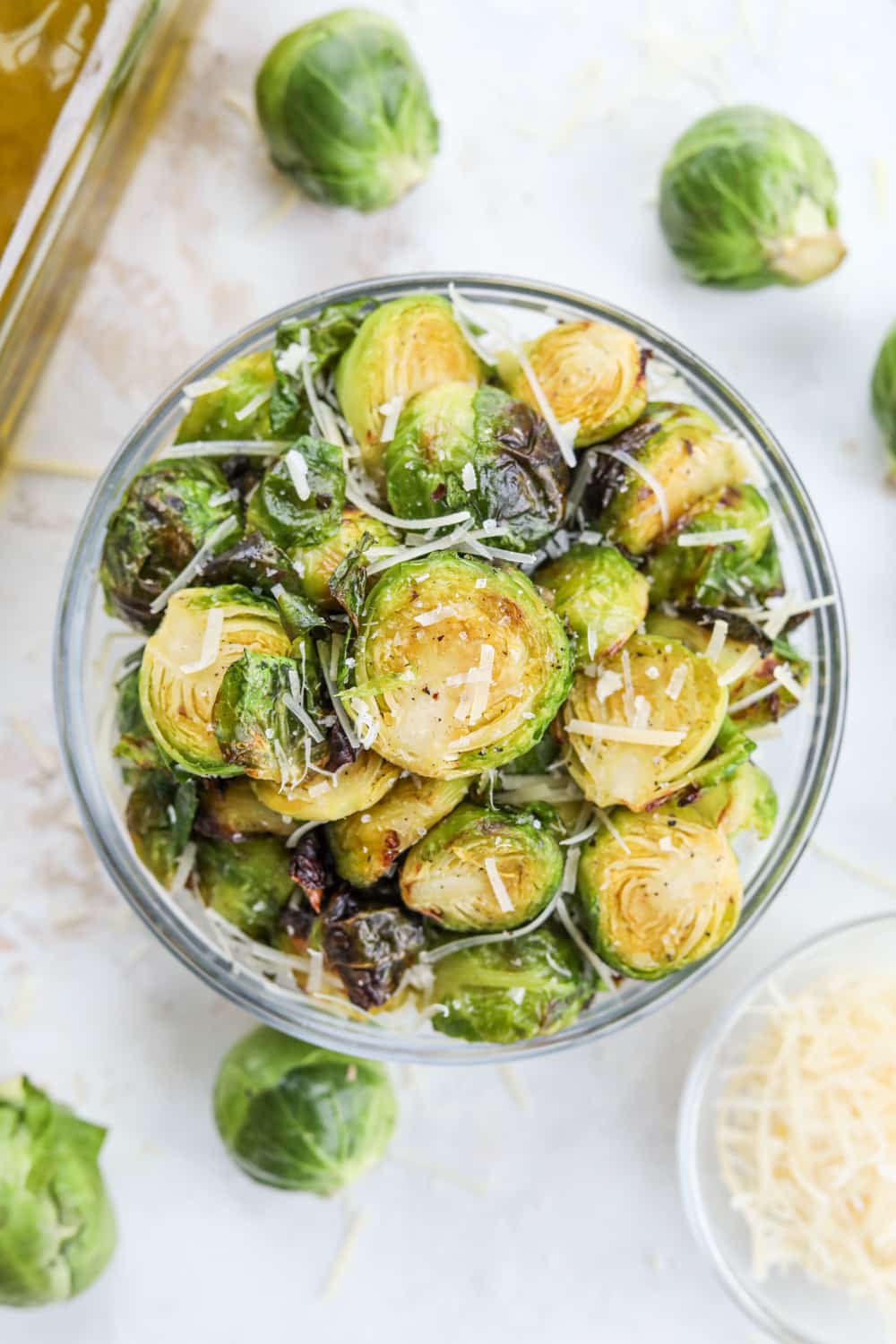 Roasted brussels sprouts topped with shredded Parmesan cheese and flaky salt in a clear glass bowl. There is a bottle of olive oil and uncooked brussels sprouts next to the bowl.