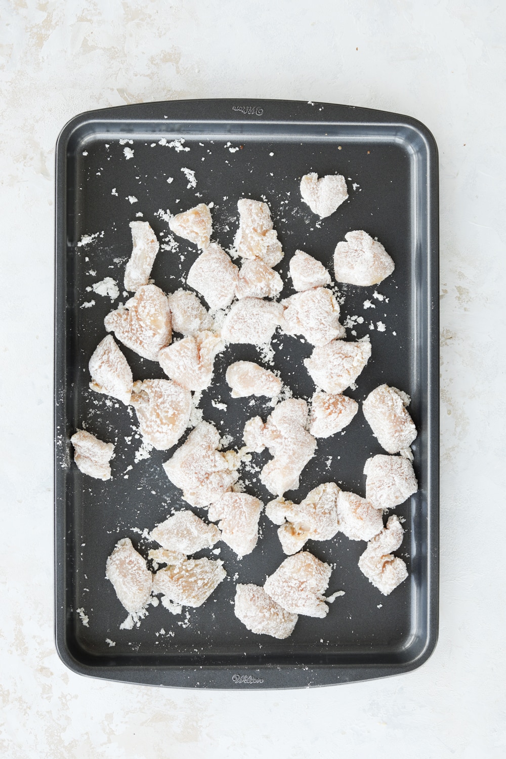 Pieces of cubed up chicken covered in unflavored protein powder on a gray baking sheet.