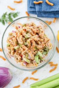 Tuna salad in a bowl. There is a stalk of celery, a red onion, a blue napkin, macaroni, and dill surrounding the bowl.