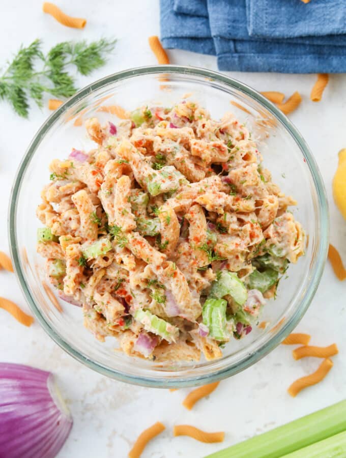 Tuna salad in a bowl. There is a stalk of celery, a red onion, a blue napkin, macaroni, and dill surrounding the bowl.