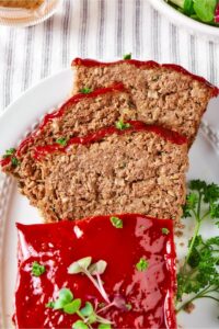 Part of a whole meatloaf with three slices overlapping one another in front of it on a white plate.