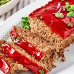 Three slices of meatloaf lying on one another touching part of a whole meatloaf on a white plate.