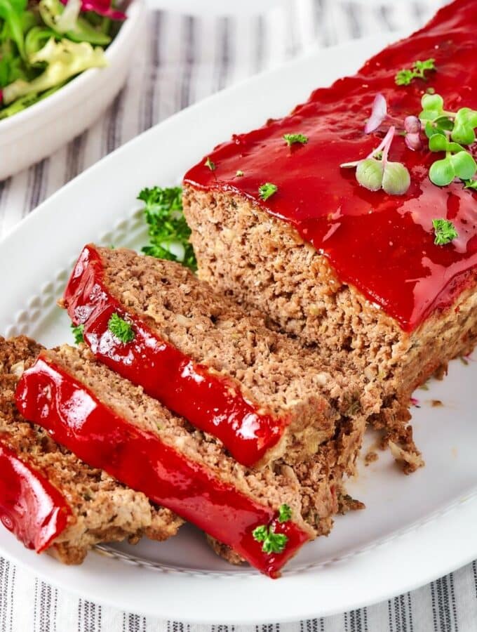 Three slices of meatloaf lying on one another touching part of a whole meatloaf on a white plate.