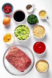 Meatloaf in a glass bowl, Parmesan cheese in a bowl, catch up in a bowl, chopped leeks in a bowl, mayonnaise and mustard in a bowl, almond flour in a bowl, soy sauce in a bowl, tomato paste in a bowl, salt and pepper in a bowl, and an egg all on a white counter.