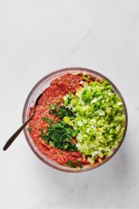 Chopped leaks and parsley on top of meatloaf mixture in a glass bowl with a spoon in it.