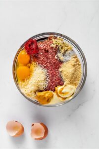 Grated Parmesan cheese, two eggs, tomato paste, almond flour, mayo, and mustard surrounding ground beef in a glass bowl on top of the white counter. In front of the bowl is a cracked egg shell.