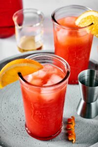 Two glasses filled with sex on the beach with an orange slice on the rim of the glass on a circular gray serving plate.