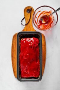 Ketchup glaze on top of meatloaf in a loaf pan that is on a wooden cutting board on a white counter. In front of the cutting board is a glass cup with a spoon in it and some ketchup smears.