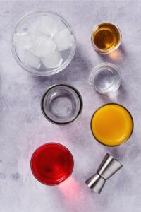 A cup of ice, a small glass pitcher of peach schnapps, a small glass pitcher of vodka, glass, a glass filled with cranberry juice, and a glass filled with orange juice on a white counter.