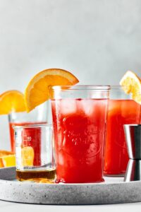 A gray serving plate with a glass filled with sex on the beach, a beaker filled with liquor behind that, another glass of sex on the beach behind that.