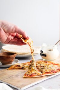 A hand pulling a slice of tortilla pizza away from the whole pizza that is on a wooden cutting board.