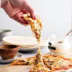 A hand holding a slice of tortilla pizza being pulled away from the whole pizza that is on a wooden cutting board.