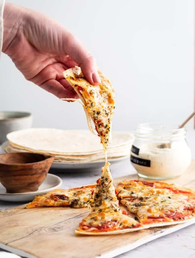 A hand holding a slice of tortilla pizza being pulled away from the whole pizza that is on a wooden cutting board.