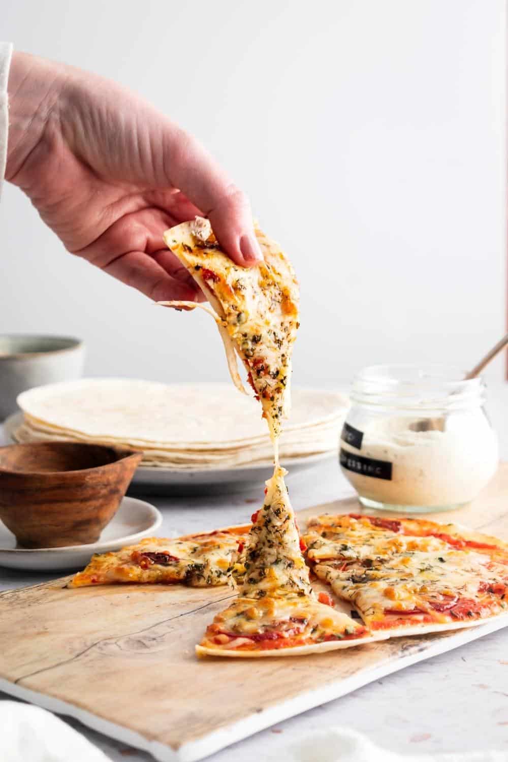 A hand holding a slice of tortilla pizza being pulled away from the whole pizza that is on a wooden cutting board.