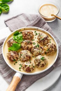 A white plate with a wooden handle that has 10 gluten-free meatballs and a white gravy covering the plate. The plate is on a tablecloth and behind the tablecloth is a glass bowl filled with the gravy.