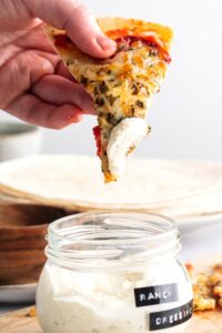 A hand holding a slice of air fryer tortilla pizza with some ranch dressing on the tip of it over a bowl of ranch dressing.