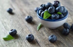 A bunch of blueberries on a wood surface surrounding a small bowl filled with blueberries.