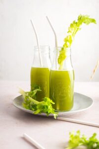 Two glasses filled with celery juice with straws in them. The glasses are on a white plate on a white counter.