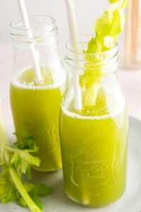 Two glasses filled with celery juice. One of the Glasses has a celery stock and a straw in it and behind that the other glass has a straw in it.