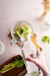 A hand pouring water into a blender that is filled with cut up celery pieces.