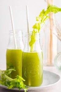 To Glasses filled with celery juice with straws in them. The glasses are on a white plate on a white.