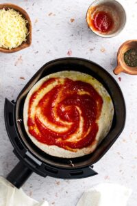 Pizza sauce on top of a tortilla in an air fryer basket.