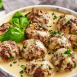 A few gluten-free meatballs that have a white gravy on top of them sitting in the white gravy that is on a white plate..