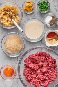 A bowl of ground beef, a bowl with an egg in it, bowl with Panko breadcrumbs, a bowl of onion, a bowl of milk, a bowl of seasoning, a bowl salt and pepper, ball cilantro, and a bowl of mustard on a gray counter.