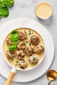 Turn gluten-free meatballs in gravy on a white plate on a white circular serving dish on a gray counter. Behind it is a glass cup with the gravy in it.