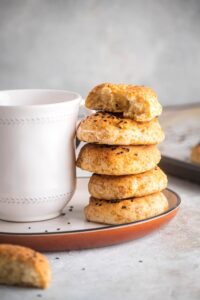 Part of a white plate with a white coffee cup on it and a stack of five bagels next to it. The top bagel is cut in half.