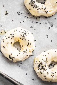 And uncooked bagel with black sesame seeds on top surrounded by parts of two other bagels on a sheet of parchment paper on a baking tray.