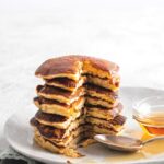 A stack of pancakes that have a piece cut out from the front of them on top of a white plate that has some syrup on it.