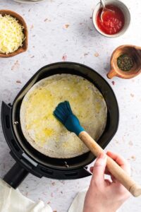 A hand holding a brush rubbing oil on a tortilla that is in an air fryer.