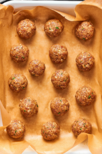 14 uncooked meat balls on a piece of parchment paper in a baking dish.