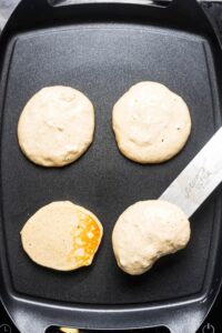 A griddle that has two circles of pancake batter in front of that is one pancake and another pancake been flipped