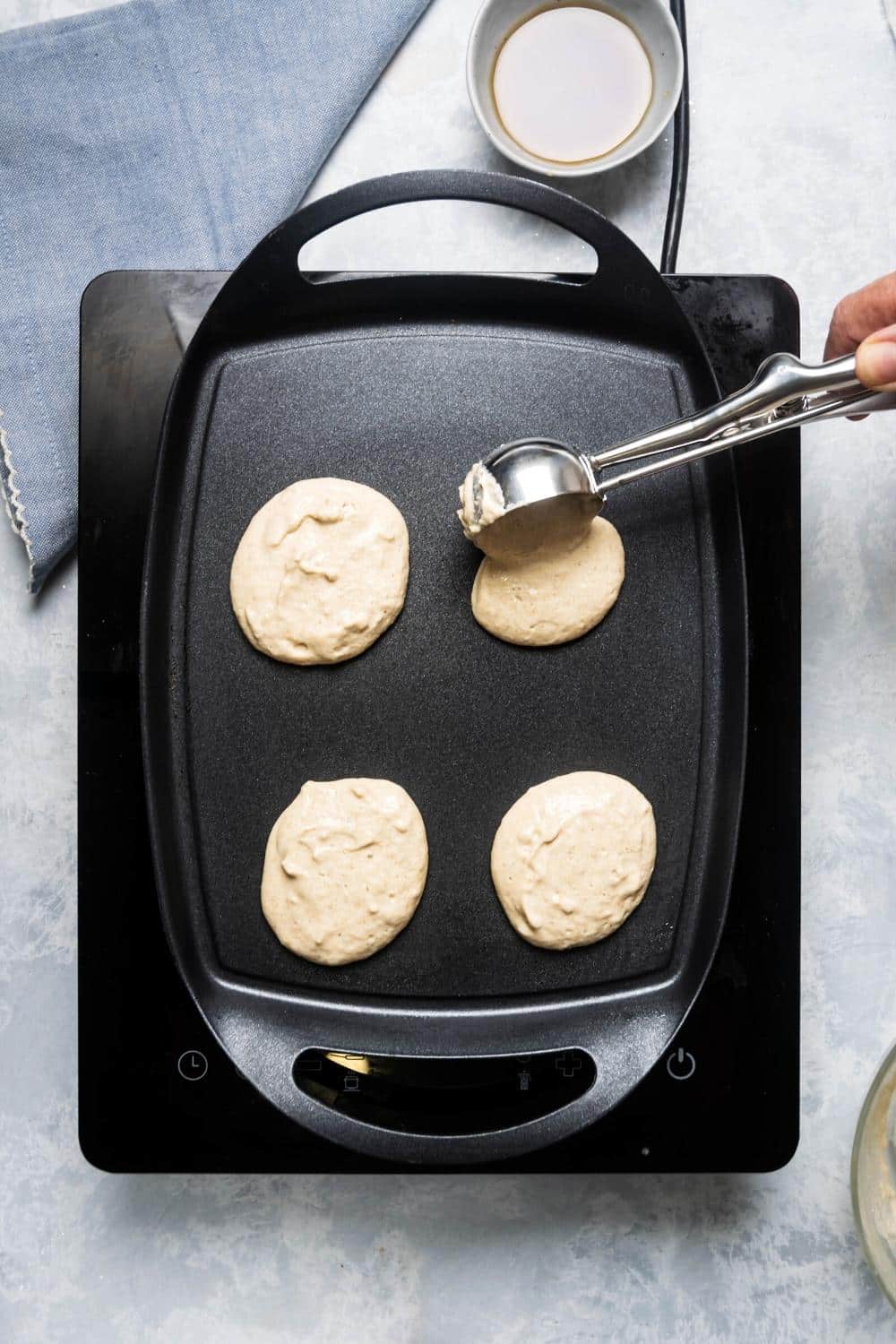 Three circles of pancake batter on a griddle and a cookie scooper pouring pancake batter onto the griddle.