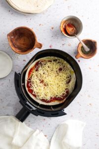 Shredded mozzarella cheese on top of pizza sauce on a tortilla in an air fryer.