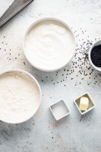 Part of a bowl of flour, a bowl of Greek yogurt, part of a bowl sesame seeds, a small square bowl of salt, and a small square bowl of butter on a white counter.