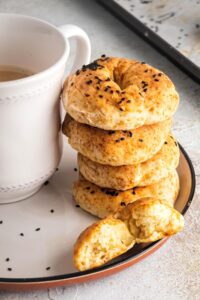 For healthy bagels stacked on top of one another on a white plate. In front of the stack is half of a bagel and next to the stack is part of a white coffee cup.