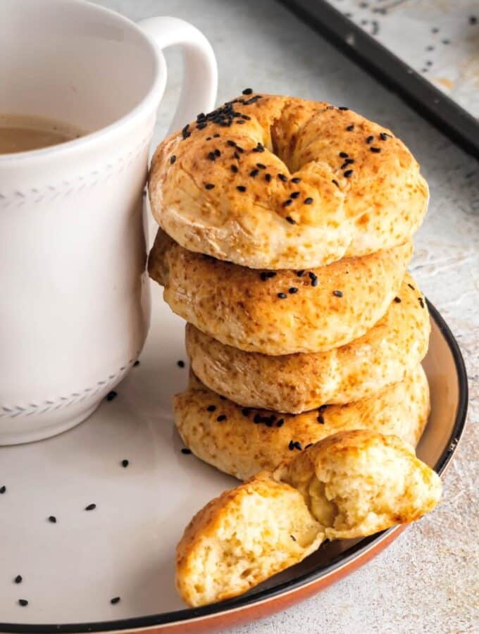 For healthy bagels stacked on top of one another on a white plate. In front of the stack is half of a bagel and next to the stack is part of a white coffee cup.