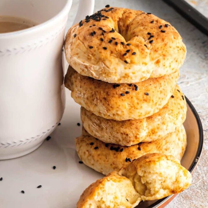 For healthy bagels stacked on top of one another on a white plate. In front of the stack is half of a bagel and next to the stack is part of a white coffee cup.